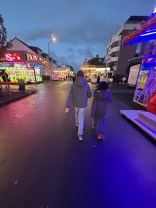 Zwei Kinder gehen am frühen Abend über eine bunte, beleuchtete Kirmesstraße. Neonlichter, Fahrgeschäfte und Regen spiegeln sich auf dem Asphalt.